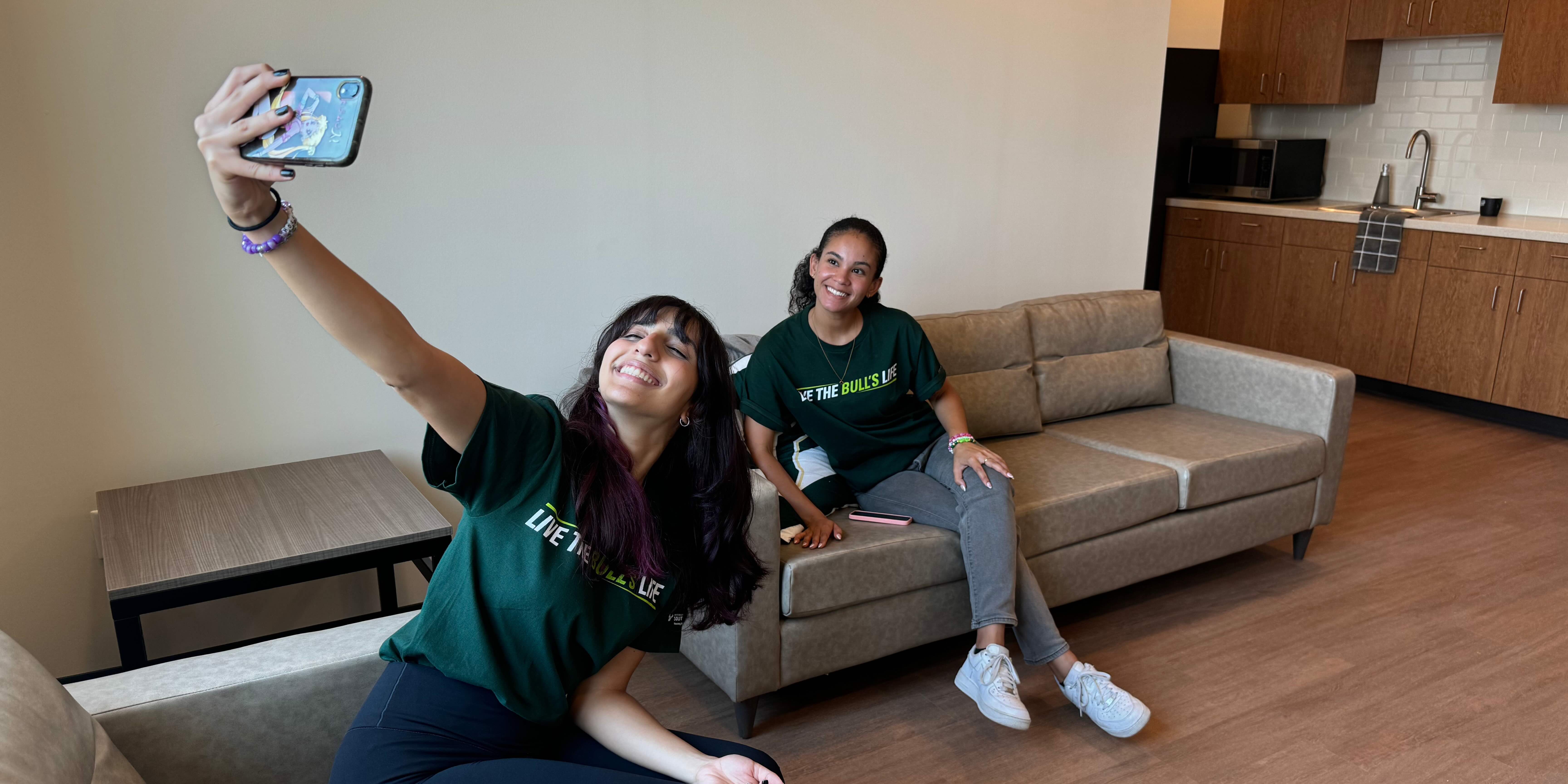 Two students in a living room. One is sitting on a couch facing the other, who has their phone in the air taking a selfie image of the two of them. 