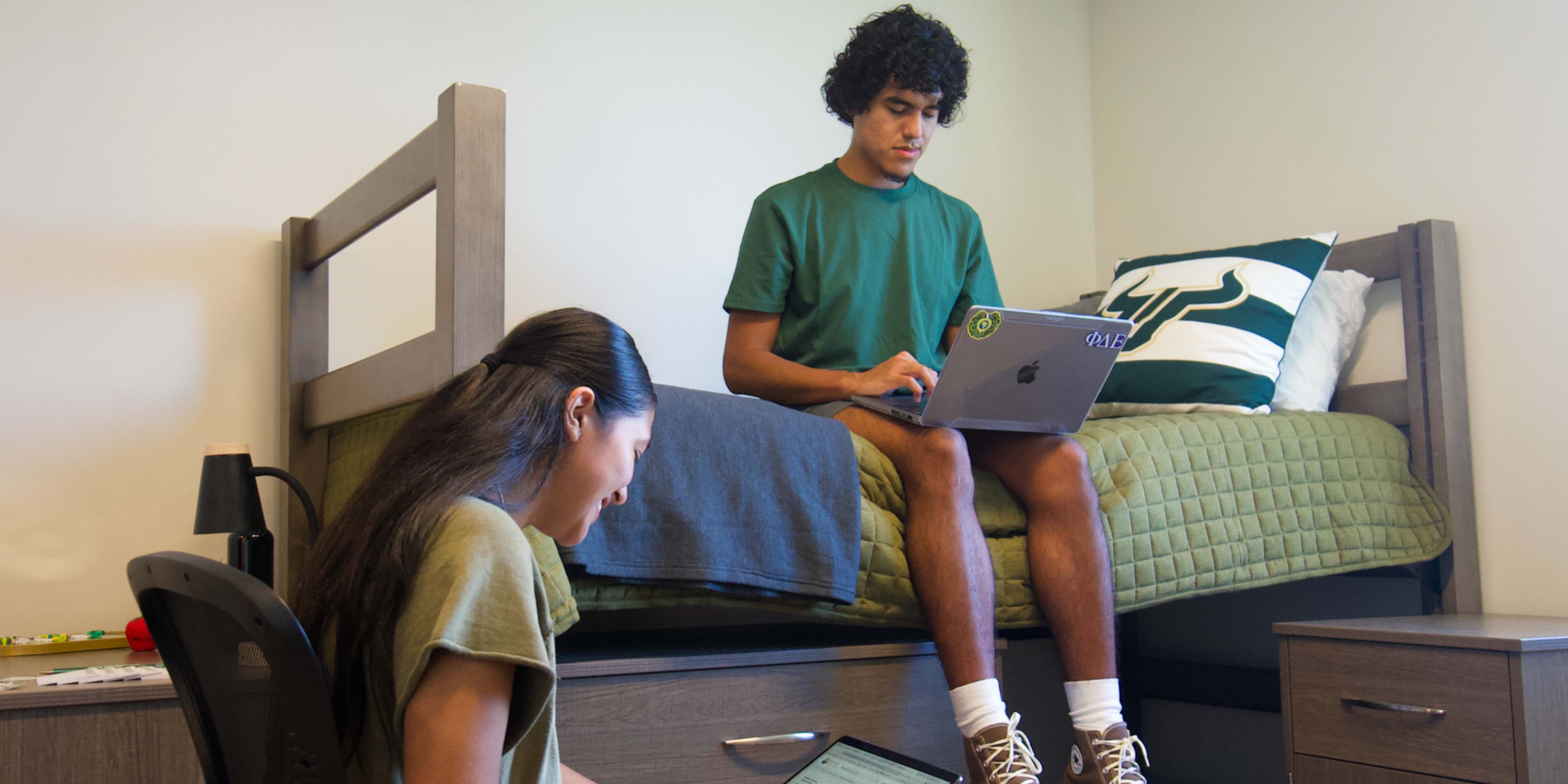 Students studying in a suite style room. A male student is on his laptop while sitting in bed and a female student is facing him in a chair with her laptop open